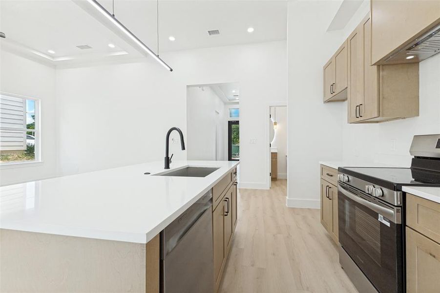 Kitchen featuring a central island with an undermount sink and matte black faucet, stainless steel appliances, light wood-finish cabinetry, and light wood-finish flooring Kitchen featuring a central island with an undermount sink and matte black faucet, stainless steel appliances, light wood-finish cabinetry, and light wood-finish flooring