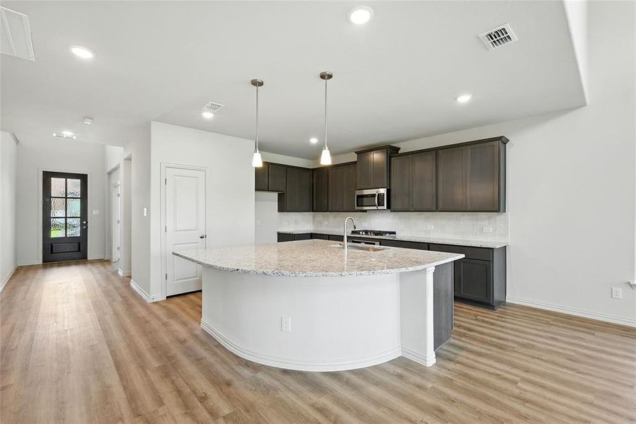 Kitchen with light stone counters, stainless steel microwave, a kitchen island with sink, backsplash, and light wood finished floors Kitchen with light stone counters, stainless steel microwave, a kitchen island with sink, backsplash, and light wood finished floors