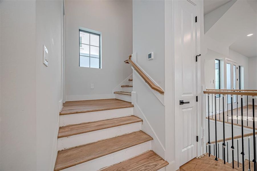 Bright and modern staircase area with light wood steps, white walls, and sleek black railings. Natural light fills the space from a window and glass doors, creating an inviting ambiance.