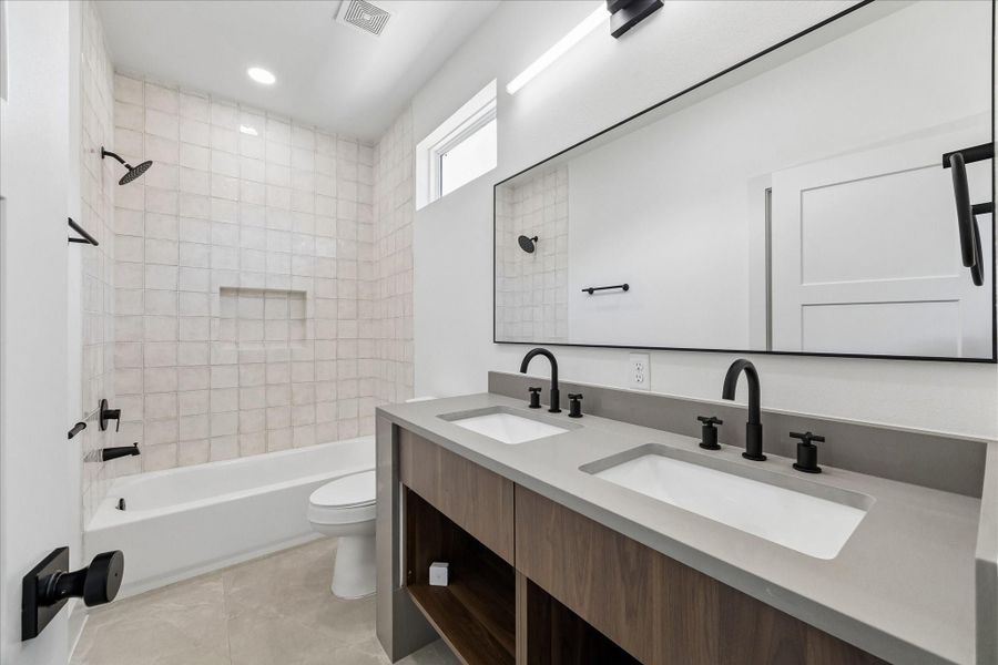 Beautifully appointed secondary bathroom features a dual sink vanity with quartz countertops, contemporary wood cabinetry, and striking matte black fixtures. A full width mirror and modern lighting enhance the bright, open feel, while the tub and shower combination showcases floor to ceiling designer tile and a built in niche for added convenience. Thoughtfully designed with upscale finishes, this bath comfortably serves multiple bedrooms while maintaining the home’s elevated modern aesthetic.