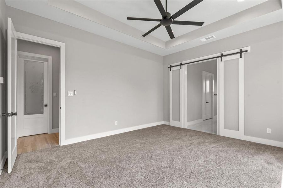 Unfurnished bedroom featuring ceiling fan, a barn door, a raised ceiling, and light colored carpet