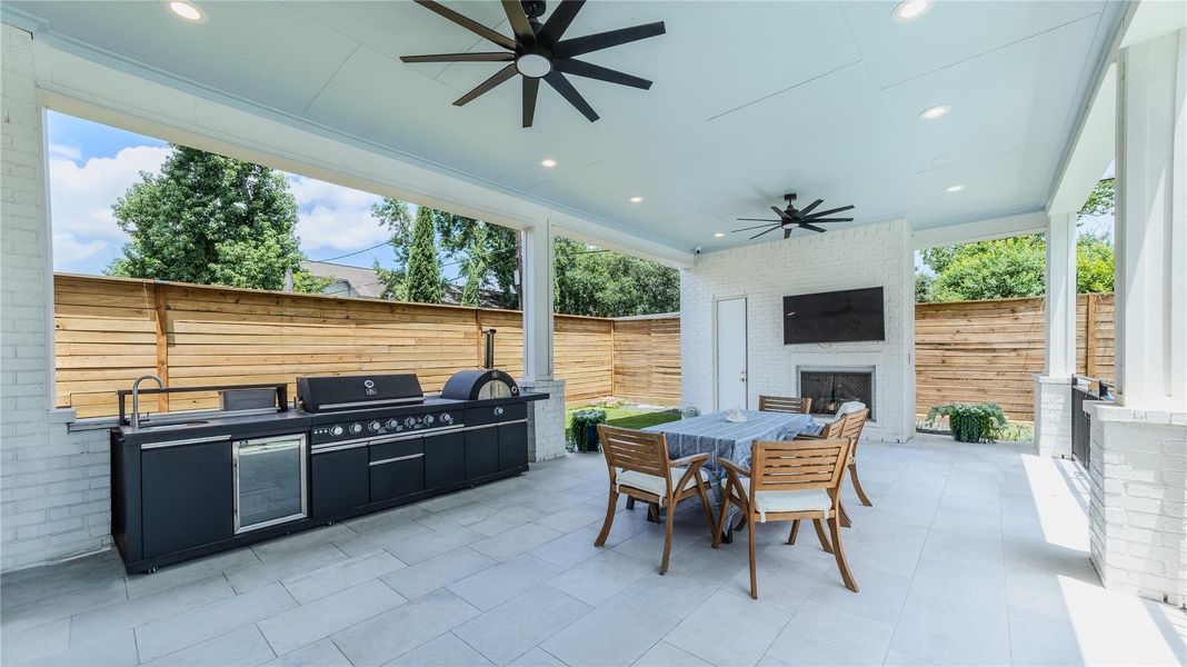 This photo showcases a spacious covered patio with a built-in outdoor kitchen, large grill, and a fireplace. It features a dining area, ceiling fans, and a wall-mounted TV, perfect for entertaining. The space is enclosed by a wooden privacy fence and surrounded by greenery.