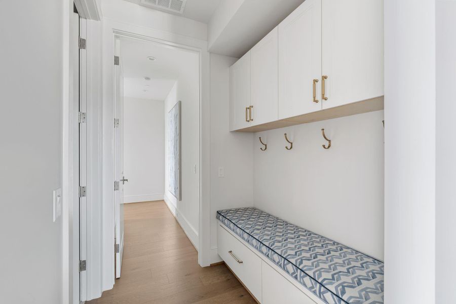 This photo shows a bright, modern mudroom with built-in white cabinetry, gold hardware, and a patterned cushioned bench. There are hooks for hanging items and light wood flooring that adds warmth to the space. This photo shows a bright, modern mudroom with built-in white cabinetry, gold hardware, and a patterned cushioned bench. There are hooks for hanging items and light wood flooring that adds warmth to the space.