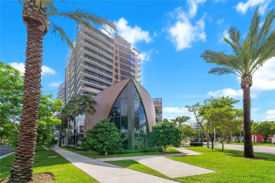 Multi-story architectural exterior featuring a curved metallic-toned facade, floor-to-ceiling glass paneling, tiered balconies, and manicured grounds with palm trees