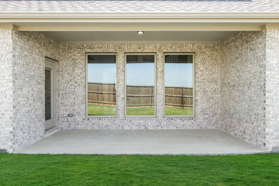 Covered back patio with concrete floor, light brick walls, and three large windows overlooking green lawn Covered back patio with concrete floor, light brick walls, and three large windows overlooking green lawn