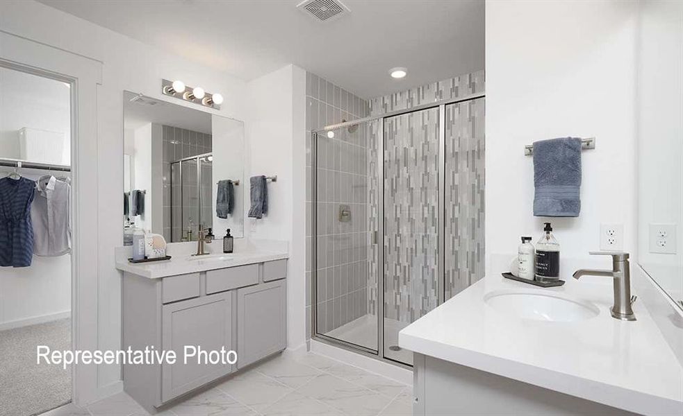 Bathroom featuring dual vanity sinks with light gray cabinetry and white countertops, a walk-in shower with tiled walls and glass enclosure, and large format tile flooring