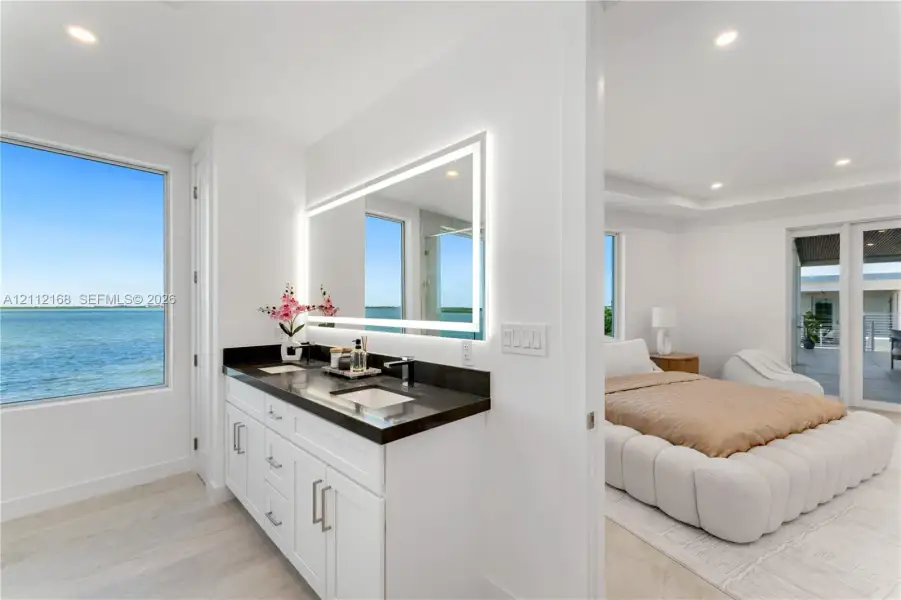 Bathroom vanity featuring white cabinetry, a dark countertop, integrated dual sinks, and an illuminated rectangular mirror, complemented by water-front exterior exposure Bathroom vanity featuring white cabinetry, a dark countertop, integrated dual sinks, and an illuminated rectangular mirror, complemented by water-front exterior exposure