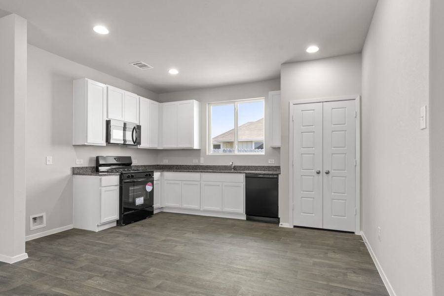 An image of kitchen with wood vinyl flooring, white cabinets with grey countertops, black appliances, a pantry, and cream colored painted walls.