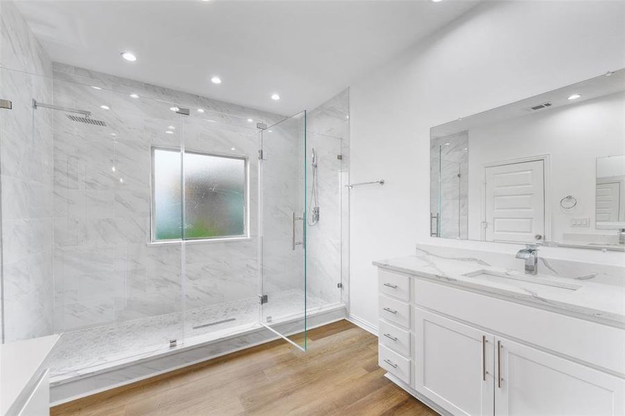 Spacious bathroom featuring a frameless glass shower enclosure with rain shower head, marble-patterned tile, a privacy window, a single vanity with stone countertop, and wood-finish flooring