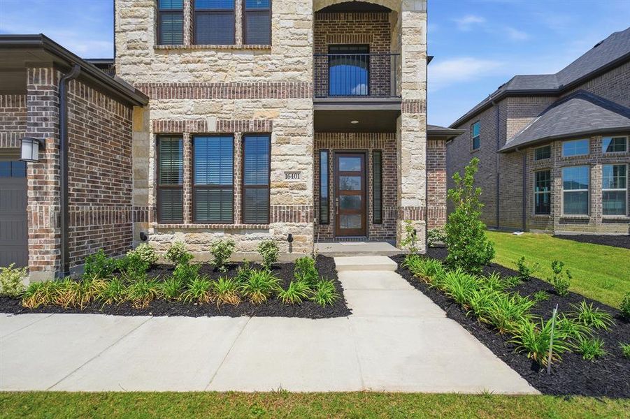 Stone and brick facade featuring a covered entry, an upper-level balcony with iron railing, a contemporary glass-paneled front door, and manicured landscaping Stone and brick facade featuring a covered entry, an upper-level balcony with iron railing, a contemporary glass-paneled front door, and manicured landscaping