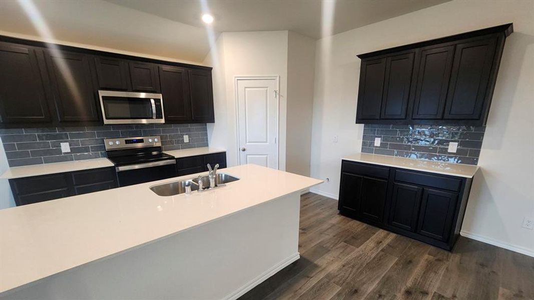 Kitchen featuring a central island with an undermount sink, dark cabinetry, light countertops, subway tile backsplash, stainless steel appliances, and wood-finish flooring