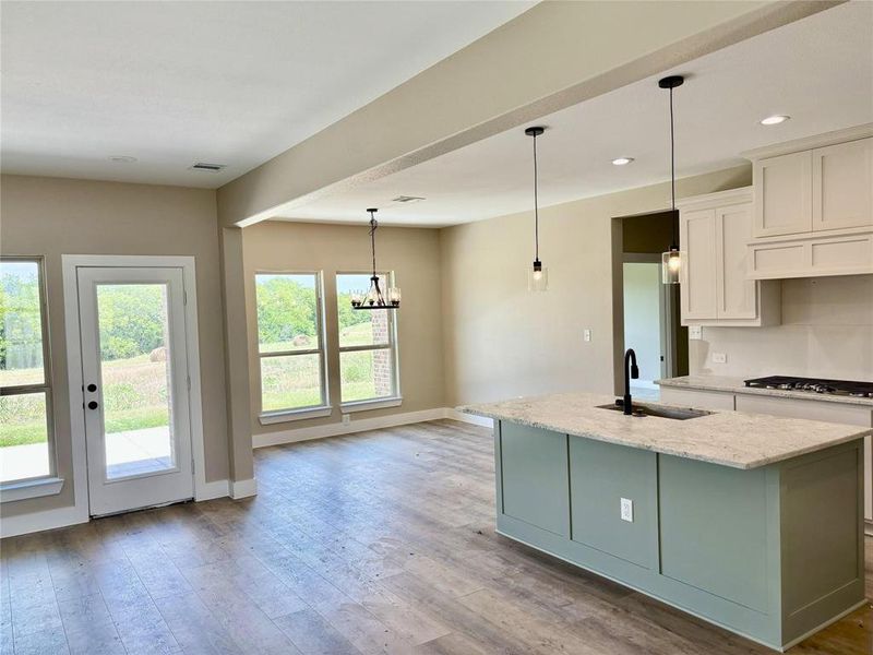 Kitchen with light wood-type flooring, light stone countertops, suspended lighting, an island with sink, and dual tone cabinetry