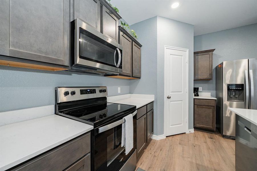 Kitchen with appliances with stainless steel finishes, light wood finished floors, dark brown cabinetry, light stone counters, and a textured wall Kitchen with appliances with stainless steel finishes, light wood finished floors, dark brown cabinetry, light stone counters, and a textured wall