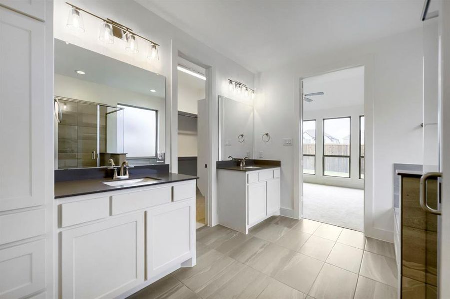 Bathroom featuring light-toned tile flooring, dual vanities with dark countertops, and white cabinetry Bathroom featuring light-toned tile flooring, dual vanities with dark countertops, and white cabinetry