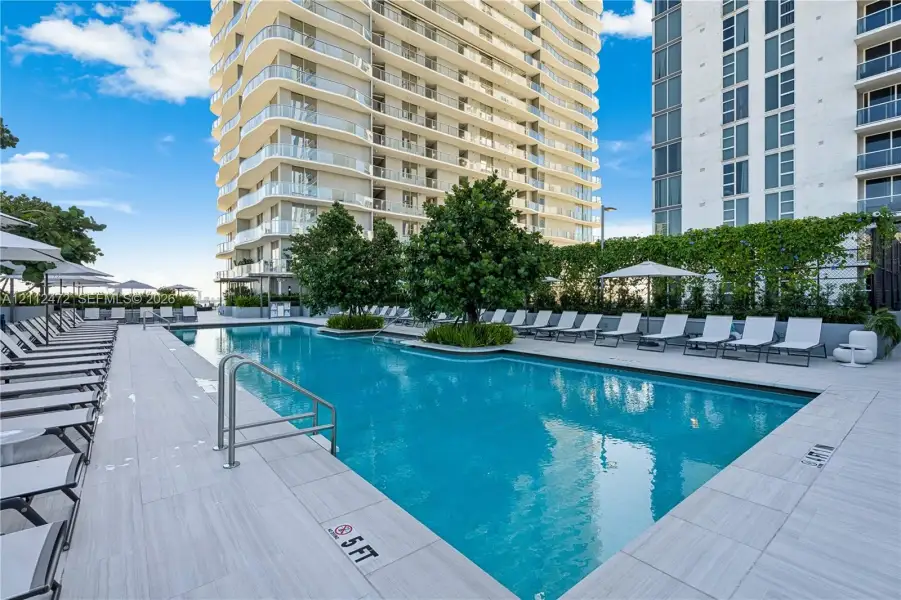 Outdoor swimming pool featuring a tile-surround deck, inset landscaping with mature trees, and metal handrails