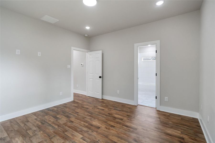 This photo shows a bright, unfurnished room with light gray walls and wood-look flooring. It features recessed lighting, and there are doors leading to a bathroom and hallway. This photo shows a bright, unfurnished room with light gray walls and wood-look flooring. It features recessed lighting, and there are doors leading to a bathroom and hallway.