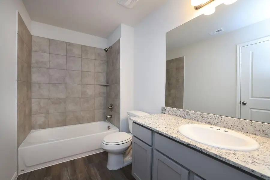 This bathroom features a modern design with a sleek granite countertop, a large mirror, and ample cabinet space. It includes a shower-tub combination, stylish tile surround and wood-look flooring, offering a clean and contemporary feel.
