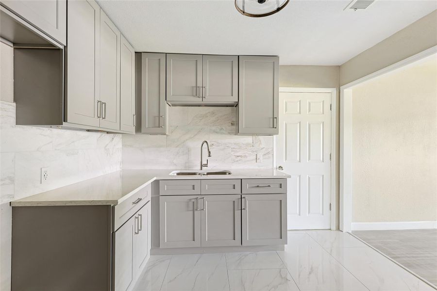 This modern kitchen features sleek gray cabinetry, a marble-style backsplash, and a clean, bright aesthetic with plenty of natural light.
