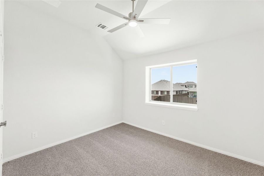 Empty bedroom with vaulted ceiling, ceiling fan, carpet flooring, and a window overlooking neighboring homes