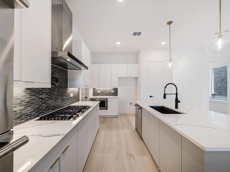 Kitchen featuring light stone counters, light wood-type flooring, wall chimney range hood, backsplash, and pendant lighting