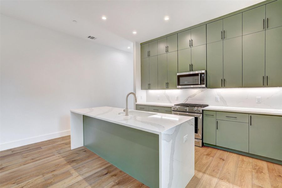 Kitchen featuring green cabinets, appliances with stainless steel finishes, backsplash, light stone counters, and light wood finished floors Kitchen featuring green cabinets, appliances with stainless steel finishes, backsplash, light stone counters, and light wood finished floors