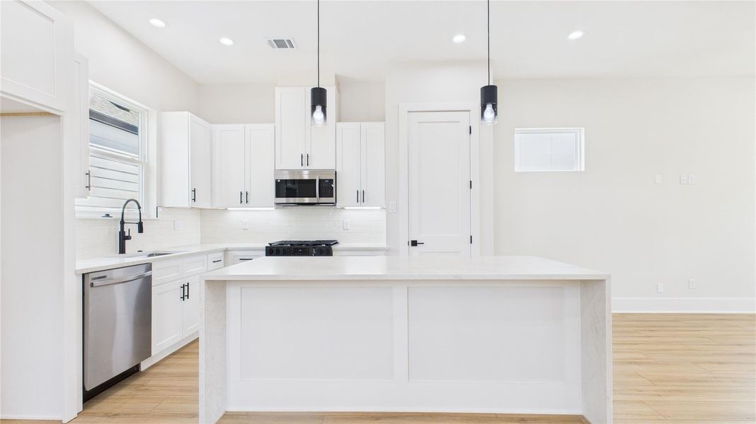 This modern kitchen features sleek white cabinetry, stainless steel appliances, and a spacious island with pendant lighting. The open layout and light wood flooring create a bright, inviting space perfect for cooking and entertaining.