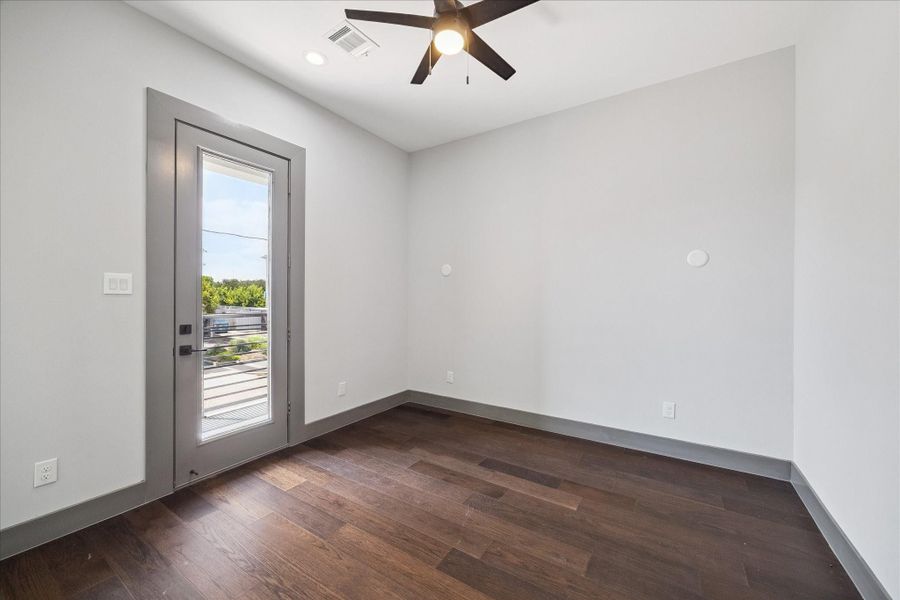 This secondary bedroom features warm hardwood flooring, neutral walls, and a ceiling fan for year-round comfort. A glass door leads directly onto a private balcony, creating an inviting space that blends indoor and outdoor living.