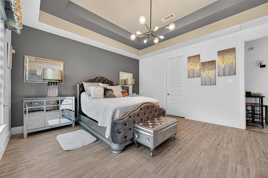 This bedroom features a modern design with a stylish tufted bed, mirrored nightstands, and a sleek chandelier. The room has a neutral color palette with a dark accent wall, decorative artwork, and wood-style flooring. A small bench sits at the foot of the bed, and the ceiling has a tray design, adding depth and elegance. This bedroom features a modern design with a stylish tufted bed, mirrored nightstands, and a sleek chandelier. The room has a neutral color palette with a dark accent wall, decorative artwork, and wood-style flooring. A small bench sits at the foot of the bed, and the ceiling has a tray design, adding depth and elegance.