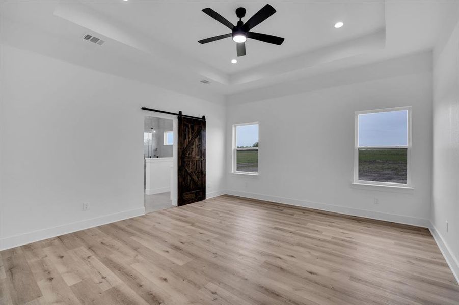 Unfurnished bedroom featuring light hardwood / wood-style floors, a barn door, ceiling fan, and a raised ceiling