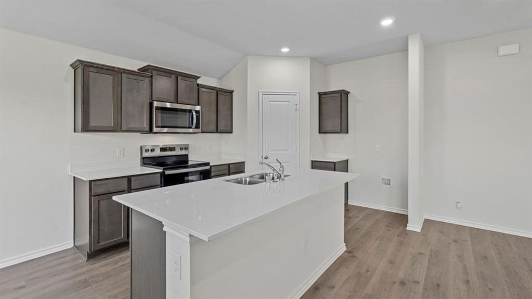 Kitchen with stainless steel appliances, dark wood finish cabinets, a center island with sink, light stone countertops, and recessed lighting
