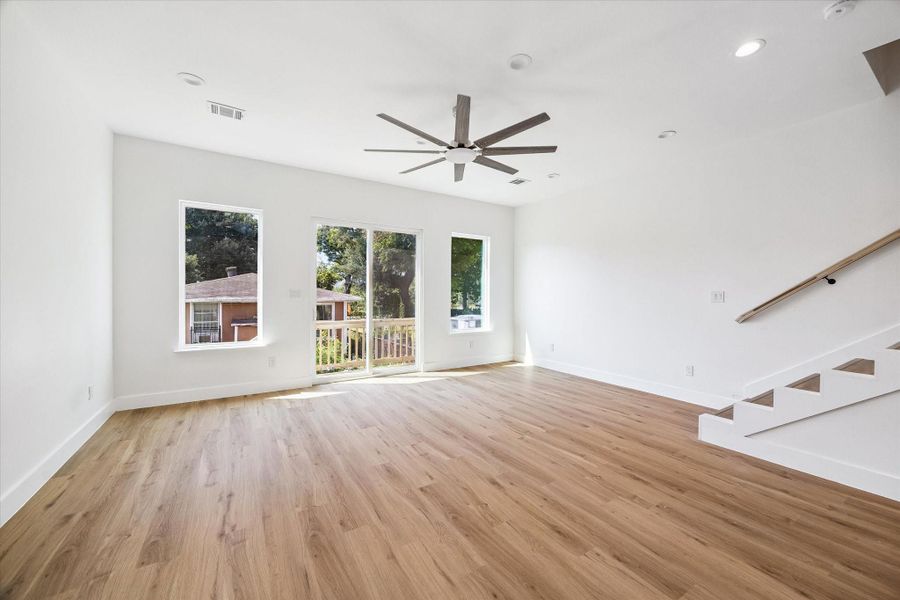 The open-concept living room offers generous space for relaxing, entertaining, or creating multiple seating zones. Large windows and sliding glass doors fill the room with natural light and provide access to the outdoor balcony. Warm wood-look flooring extends throughout the space, while recessed lighting and a contemporary ceiling fan complete the modern design. The staircase nearby creates an easy connection between levels.
