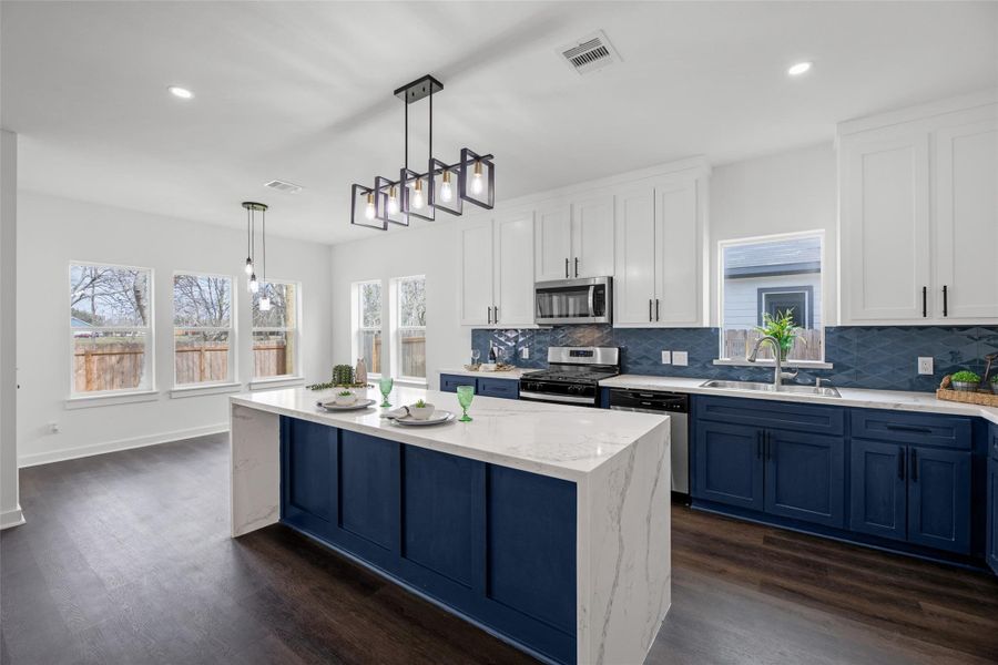 Kitchen features a spacious island with a sleek marble countertop, blue cabinetry, and stainless steel appliances. Large windows and stylish lighting fixtures provide ample natural light and a contemporary feel.