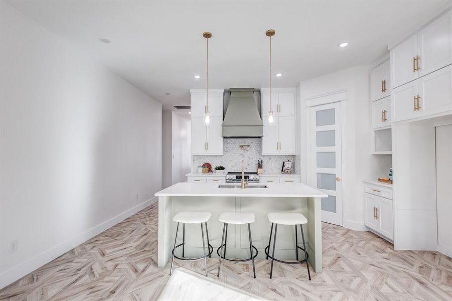 Kitchen featuring premium range hood, white cabinets, a center island with sink, sink, and hanging light fixtures