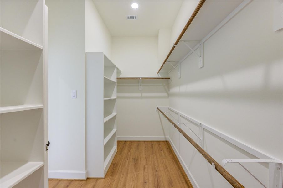 This photo shows a spacious walk-in closet with wooden flooring, ample shelving, and hanging space on both sides, providing excellent storage options. This photo shows a spacious walk-in closet with wooden flooring, ample shelving, and hanging space on both sides, providing excellent storage options.