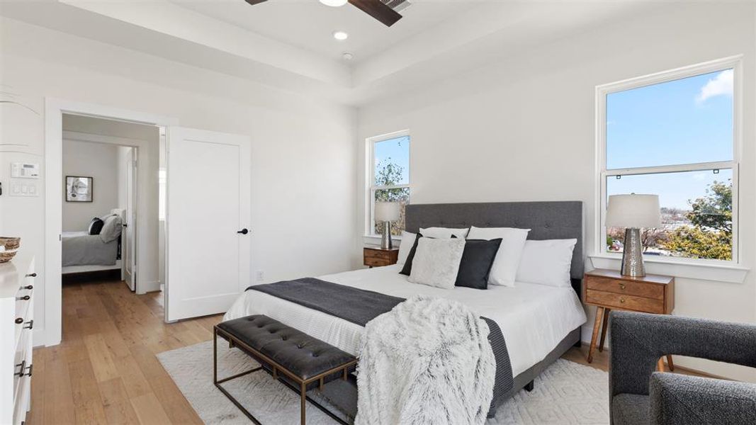 Bright and inviting bedroom featuring a comfortable bed with a gray upholstered headboard, stylish side tables with lamps, and large windows allowing natural light. The room has wooden flooring and a cozy seating area, creating a warm and modern atmosphere.