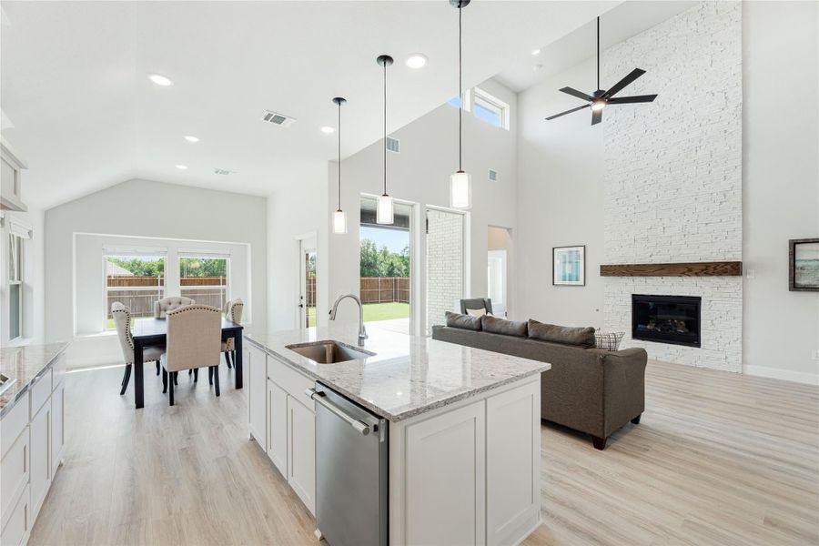 Open-concept kitchen with granite island, flowing into living room with stone fireplace and high ceilings