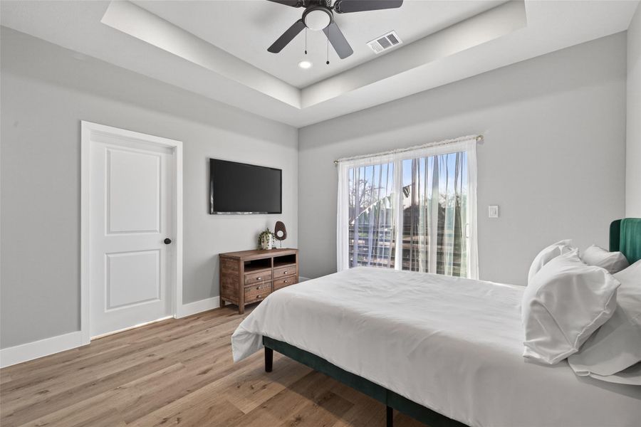 Serene primary suite featuring tray ceiling with recessed lighting, ceiling fan, white oak–finish plank laminate flooring, and private balcony access for seamless indoor-outdoor living.