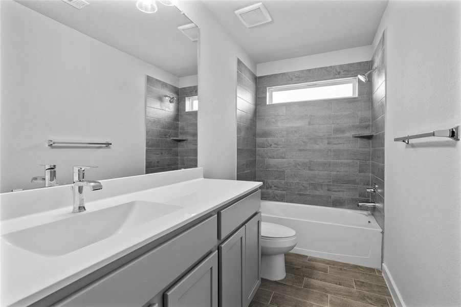 Secondary bath features tile flooring, bath/shower combo with tile surround, light stained wood cabinets, beautiful light countertops, mirror, dark, sleek fixtures and modern finishes.