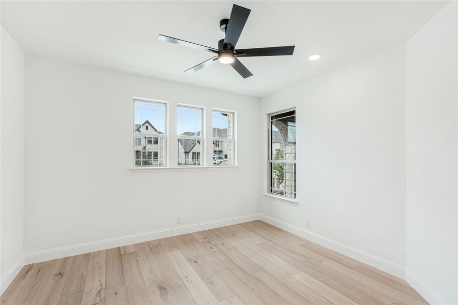 Empty bedroom with light hardwood floors, white walls, ceiling fan, and multiple windows
