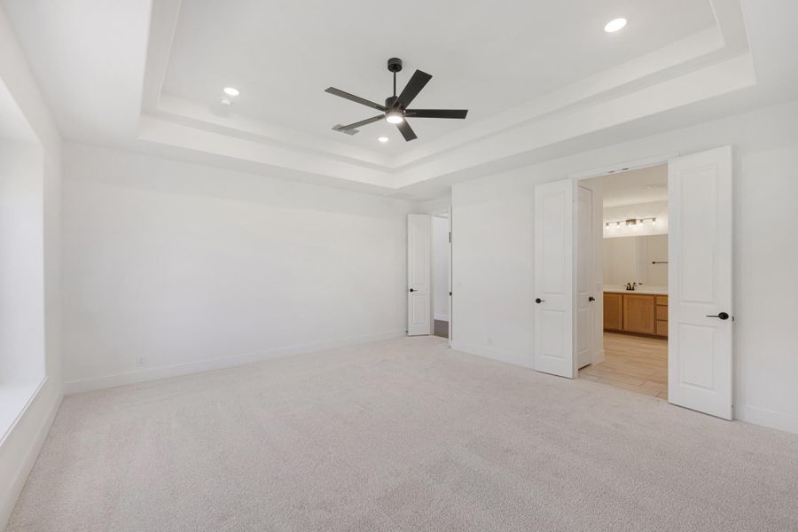 Empty bedroom with tray ceiling, ceiling fan, carpet floors, and open door to ensuite bathroom