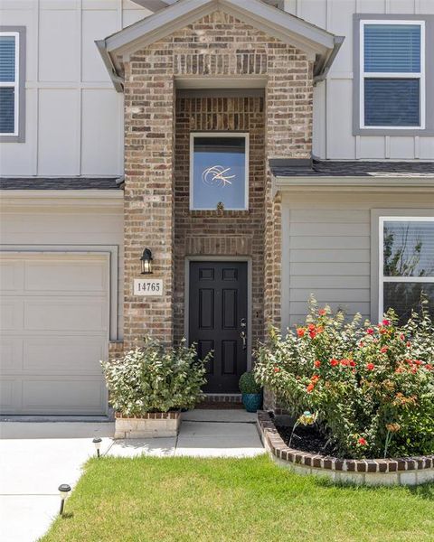 Brick and siding exterior featuring a covered entryway with a dark paneled door, an attached garage, and landscaped garden beds with mature shrubs Brick and siding exterior featuring a covered entryway with a dark paneled door, an attached garage, and landscaped garden beds with mature shrubs