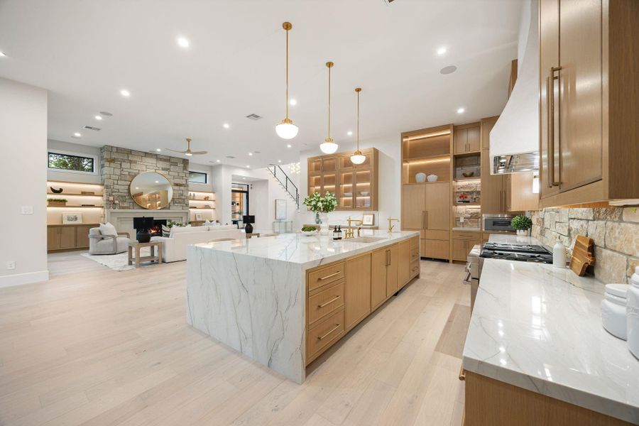 A stunning open-concept kitchen and living space designed for both everyday living and effortless entertaining. The oversized waterfall island anchors the room with clean lines and elegant stone surfaces, complemented by warm wood cabinetry and soft ambient lighting. The true standout is the seamless connection to the living area, where a floor-to-ceiling stone fireplace creates a striking focal point, blending texture, warmth, and modern sophistication.