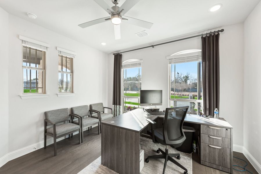 Home office with L-shaped desk, office chair, ceiling fan, and multiple windows with dark curtains
