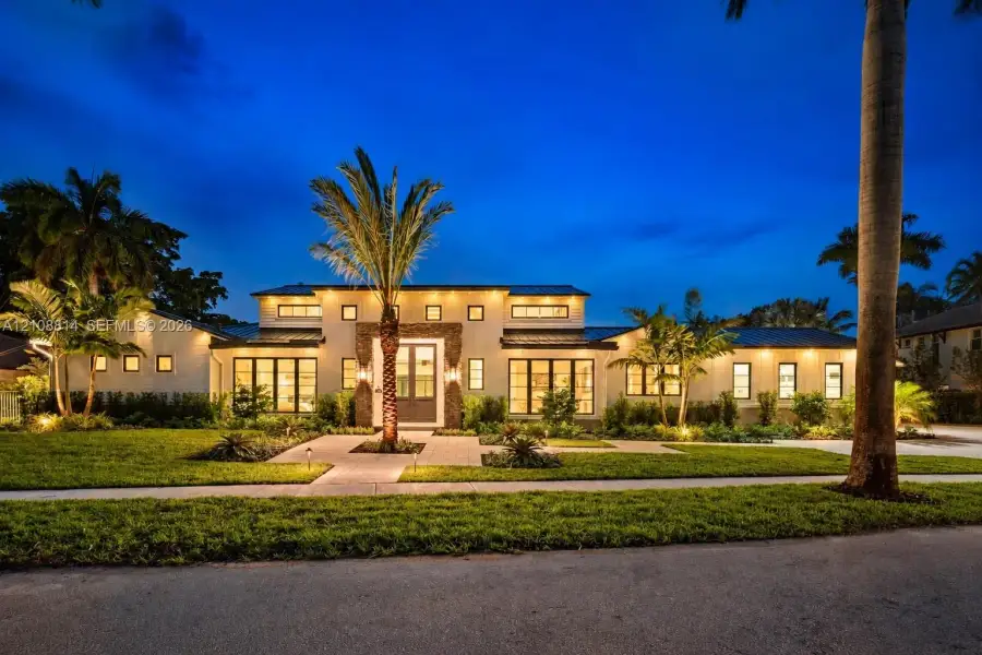 Single-story residence featuring light-toned stucco exterior, stacked stone facade accents, dark metal roof, multiple transom windows, and paved stone walkway Single-story residence featuring light-toned stucco exterior, stacked stone facade accents, dark metal roof, multiple transom windows, and paved stone walkway