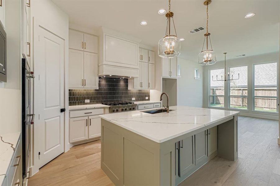 Kitchen featuring decorative backsplash, light stone counters, a center island with sink, light wood-type flooring, and decorative light fixtures Kitchen featuring decorative backsplash, light stone counters, a center island with sink, light wood-type flooring, and decorative light fixtures