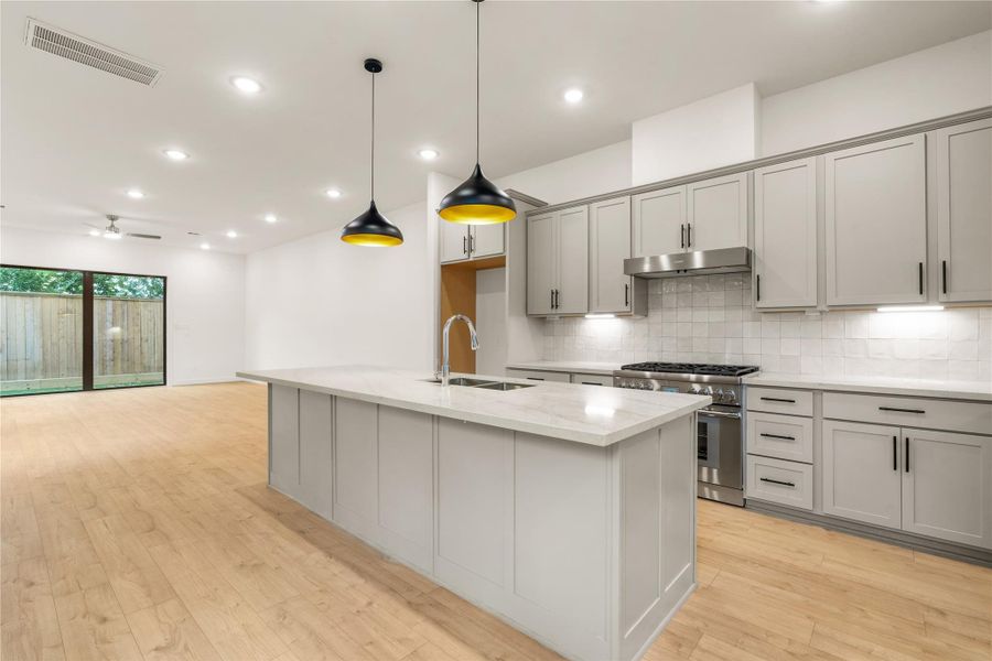 Chef's kitchen perspective — massive quartz island with bar seating potential, statement matte black and gold pendant lighting, gas cooktop setup, and soft grey shaker cabinetry extending to the main floor living space.
