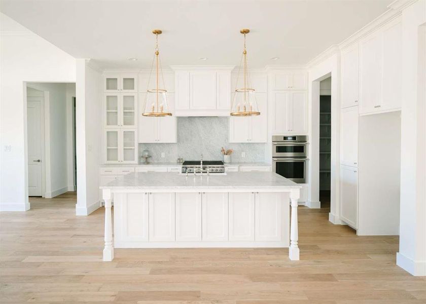 Kitchen with light wood-type flooring, white cabinetry, stainless steel double oven, a center island with sink, and decorative light fixtures