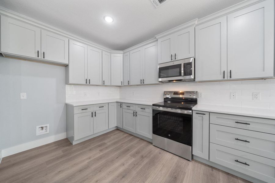 Modern kitchen featuring light gray shaker-style cabinetry with dark hardware, white subway tile backsplash, white countertops, stainless steel appliances, and wood-finish flooring