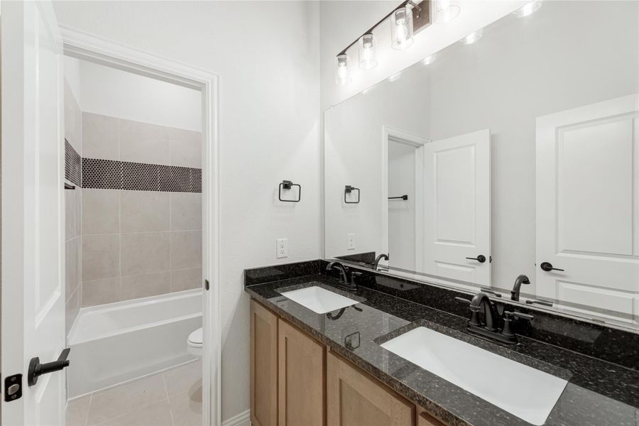 Bathroom with double sink vanity, dark granite countertop, and tiled shower-tub combo visible through doorway