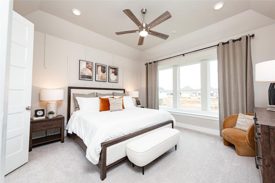 This luxurious primary suite serves as a private sanctuary, featuring a stately tray ceiling paired with a modern ceiling fan. A massive bank of windows floods the entire room with incredible natural light, while the spacious layout easily allows for versatile room arrangements.